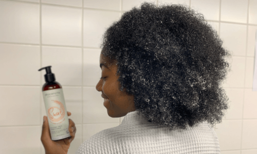 Featured image of afro haired woman with wet hair holding a bottle of Afrocenchix Swish how often should you wash your hair blog post