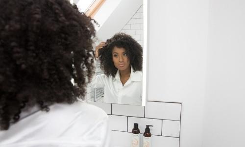 young black woman wearing white touching her hair and looking in a mirror